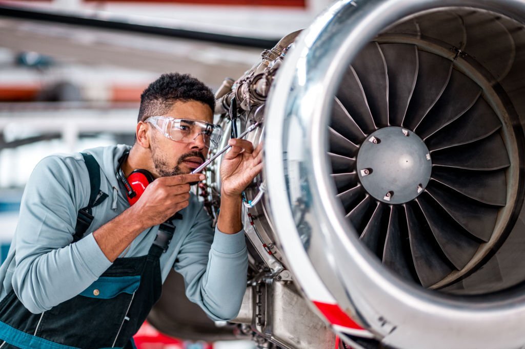 A aircraft maintenance engineer wearing glasses, ear protectors, and a utility vest, working at HAL Pravara Aviation Institute